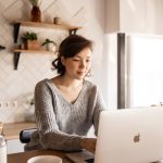 Young female in gray sweater sitting at wooden desk with laptop and bottle of milk near white bowl while browsing internet on laptop during free time at home