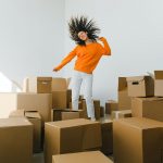 Joyful female with moving hair dancing among piles of carton containers after renting house on light background
